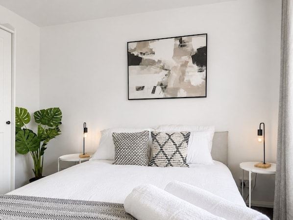 A modern bedroom with a large bed, two bedside tables, a wall-mounted abstract painting, and a potted plant.