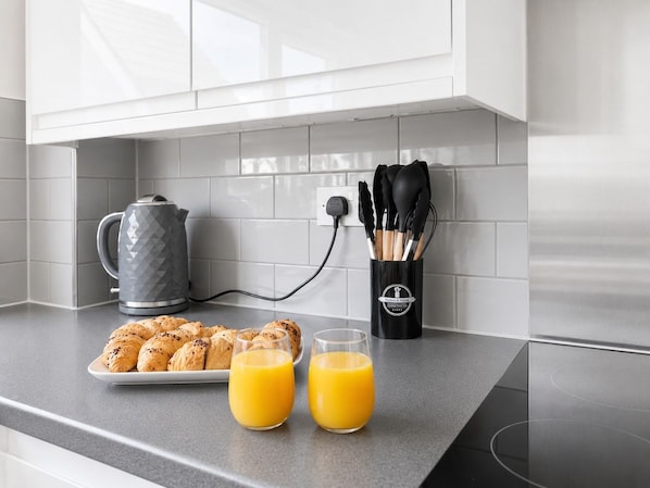 A kitchen counter with a plate of croissants, two glasses of orange juice, a kettle, and a knife holder.