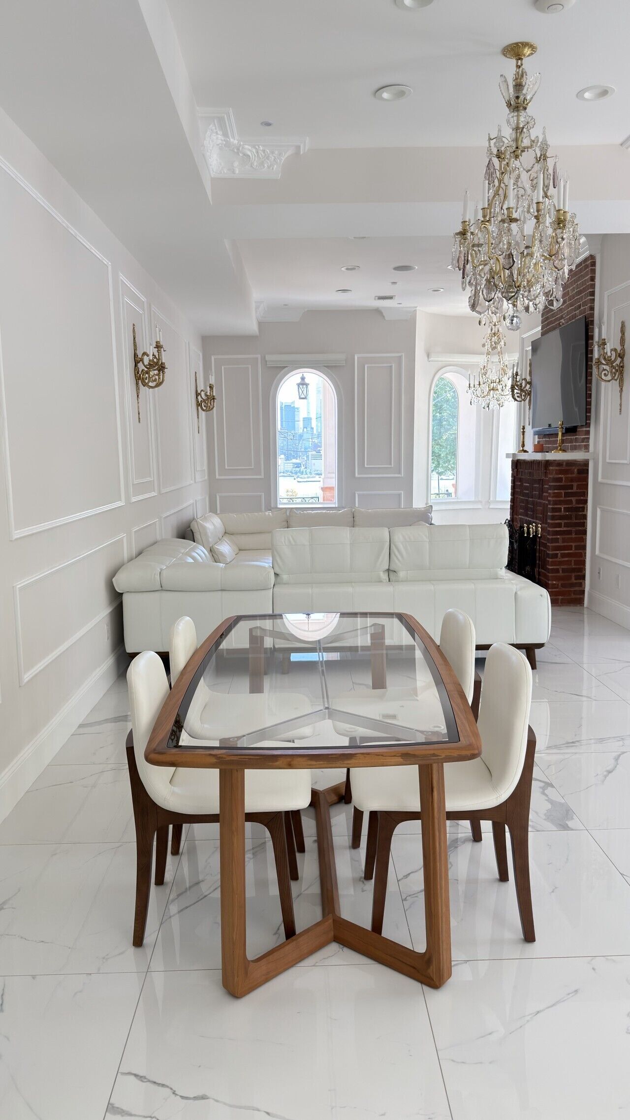 A spacious living room with a central glass-top table surrounded by chairs, a white sofa, and a chandelier.