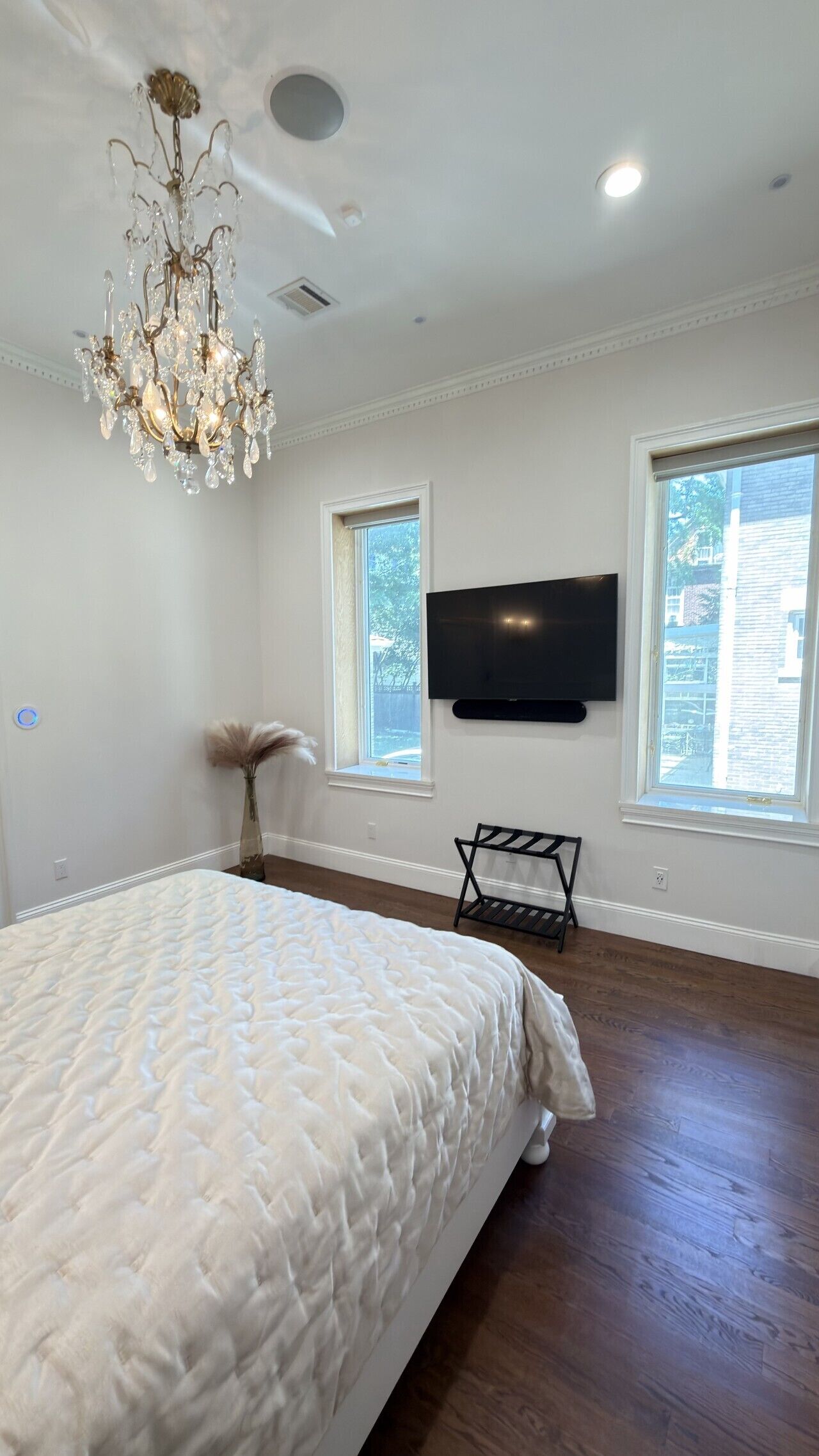 A bedroom with a large bed, a chandelier, a flat-screen TV, and two windows.