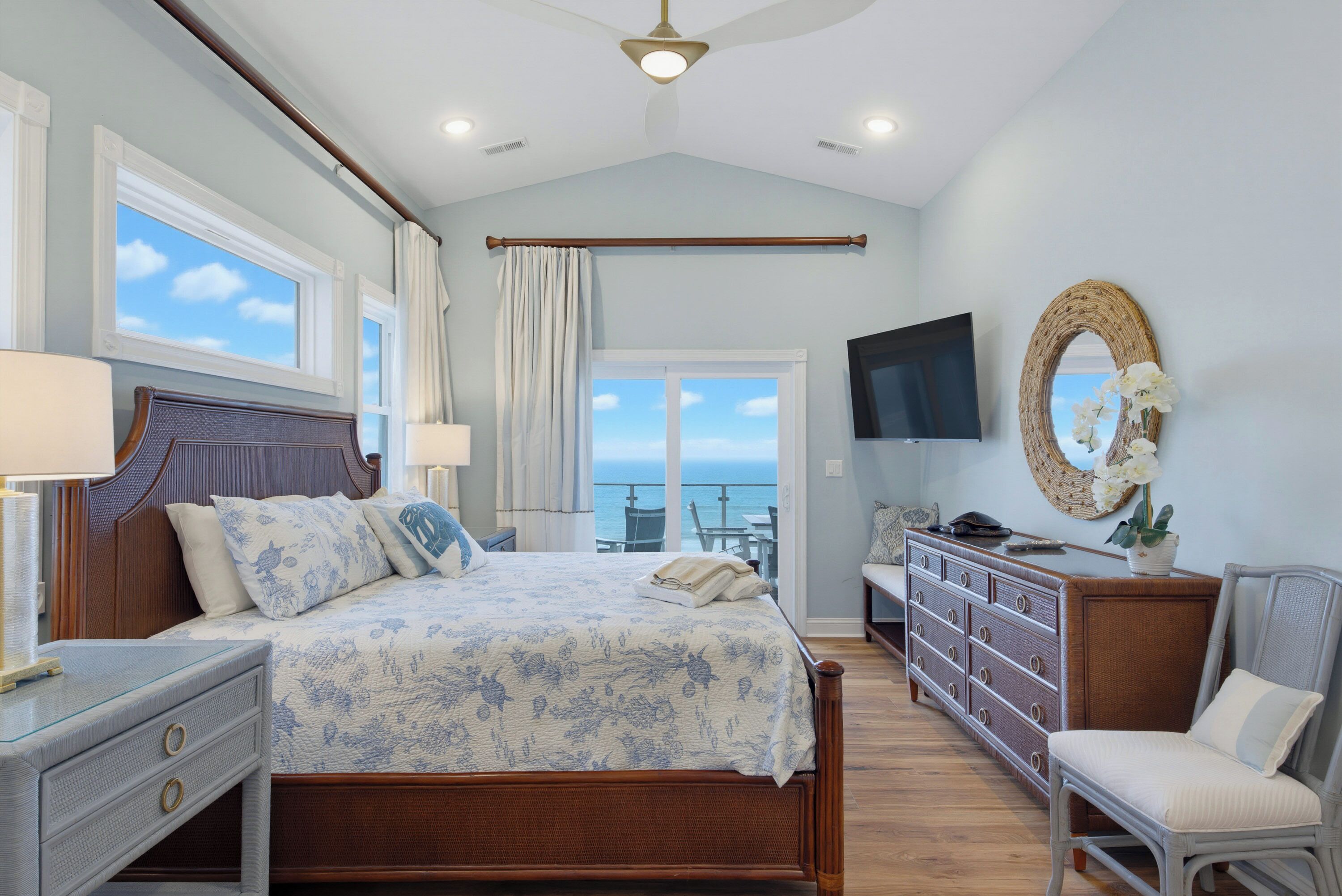 A bedroom with a bed, bedside tables, a dresser, a TV, and a view of the sea.
