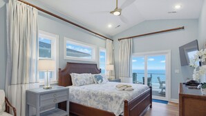 Een slaapkamer met een groot bed, twee nachtkastjes, een houten hoofdbord, een plafondventilator en uitzicht op de oceaan door een schuifdeur van glas.