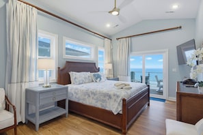 A bedroom with a large bed, two bedside tables, a nightstand, a wooden headboard, a ceiling fan, and a view of the ocean through a sliding glass door. - Oceanfront pool with theater and games (North Topsail Beach)