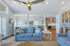 A spacious living room with a blue sofa, a dining area, and a kitchen with wooden cabinets. - Oceanfront pool with theater and games (North Topsail Beach)