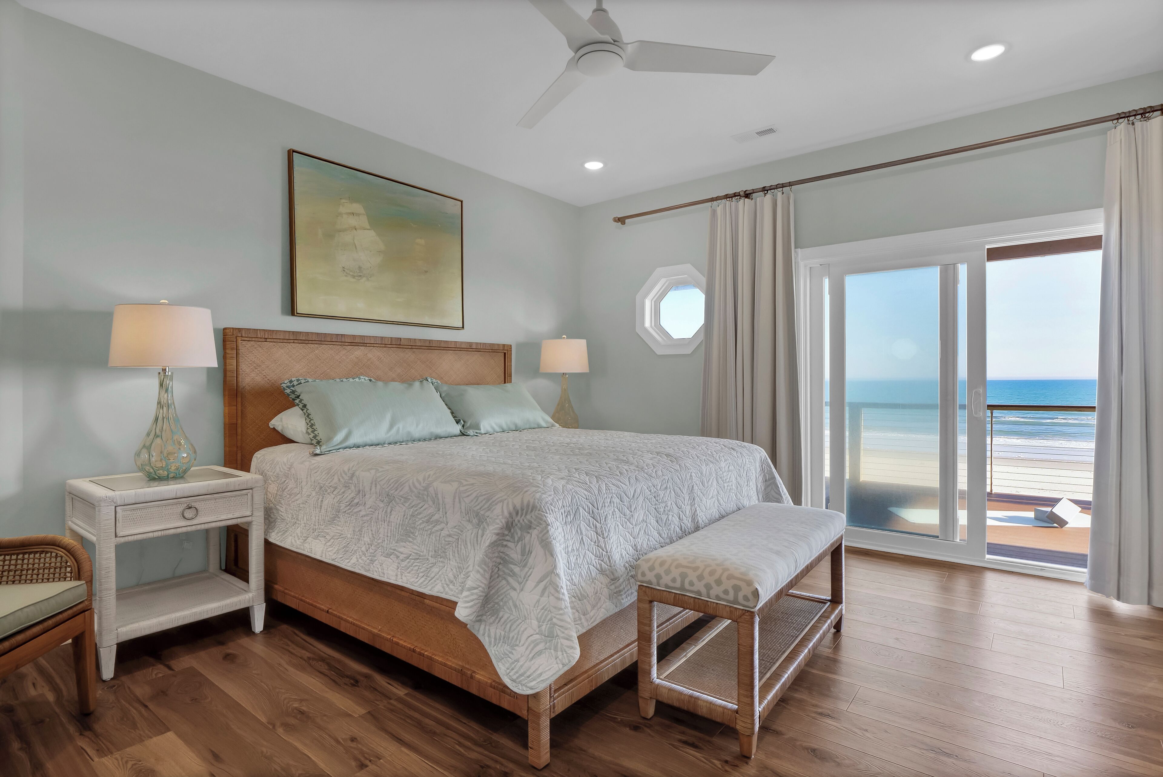 A bedroom with a bed, bedside tables, a bench, a ceiling fan, and a large window with a view of the beach.