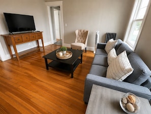 A living room with a flat-screen TV, a wooden cabinet, a grey sofa, a beige armchair, and a coffee table with decorative items.