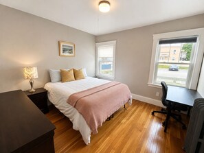 Ein Schlafzimmer mit Bett, Nachttisch, Lampe, Fenster und Schreibtisch mit Stuhl.