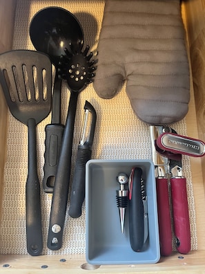 A drawer containing kitchen utensils including a spatula, a slotted spoon, a grater, a pair of tongs, and a red-handled can opener.