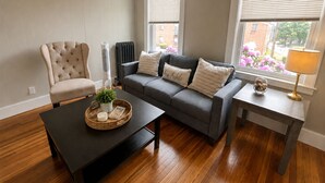 A living room with a grey sofa, a black coffee table, a beige armchair, and a window with blinds.