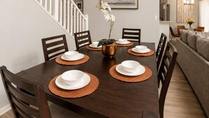 A dining room with a dark wooden table set for six, surrounded by chairs, with a staircase on the left and a living area on the right. - 5BR Fantastic Villa  Pool & Game Room Near Disney (Kissimmee)