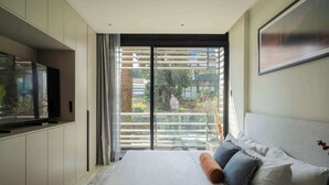 Ein modernes Schlafzimmer mit einem großen Bett, einem Flachbildfernseher und einer Glastür, die auf einen Balkon mit Blick ins Grüne führt.