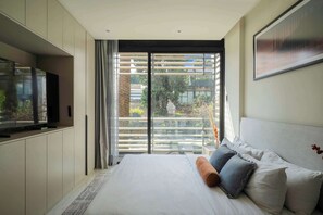 A modern bedroom with a large bed, a flat-screen TV, and a sliding glass door leading to a balcony with a view of greenery. - Family 2BR Apartment with Street-View Balcony in Polanco (Ciudad de México)