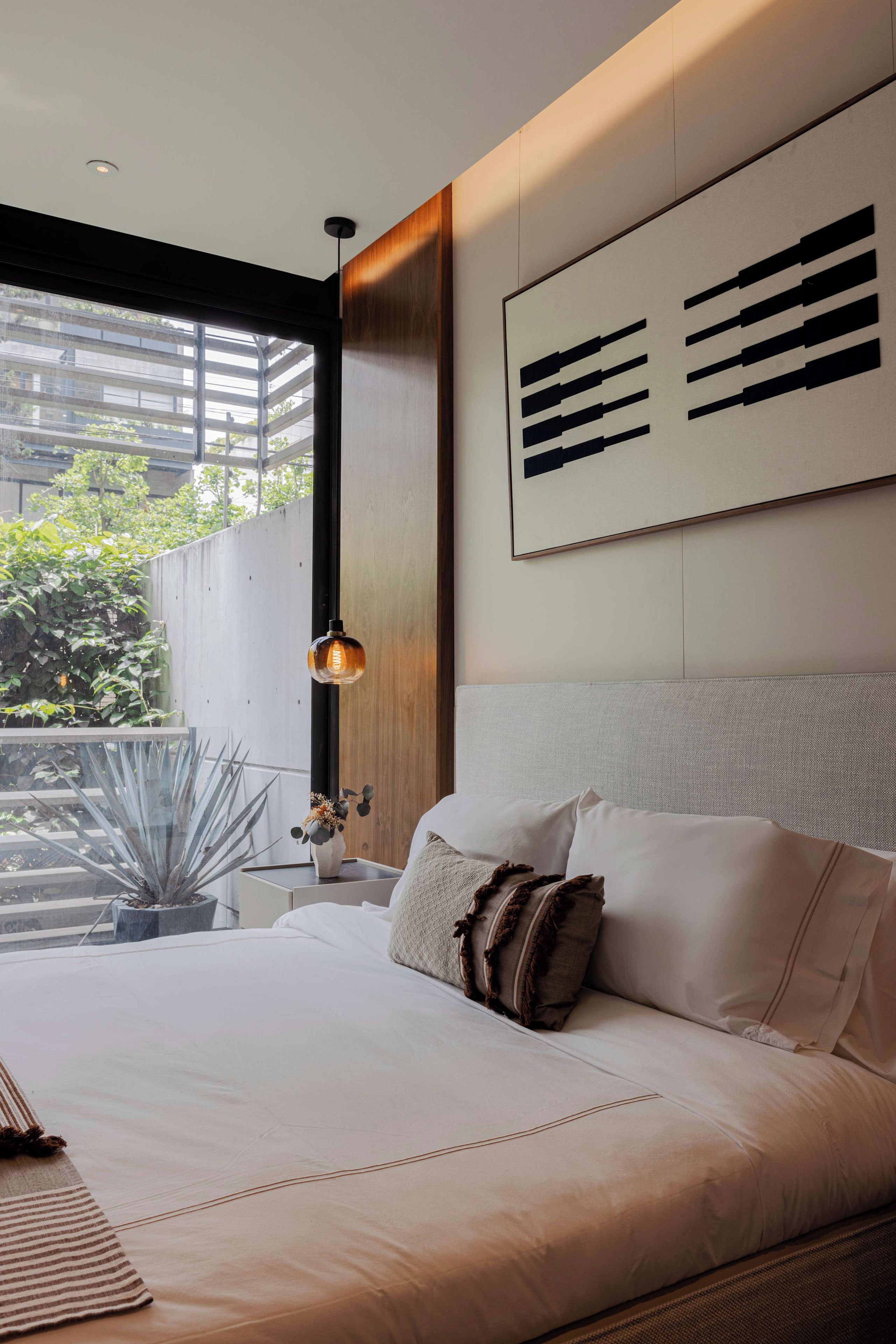 A modern bedroom with a large bed, a bedside table with a lamp, and a view of greenery outside.