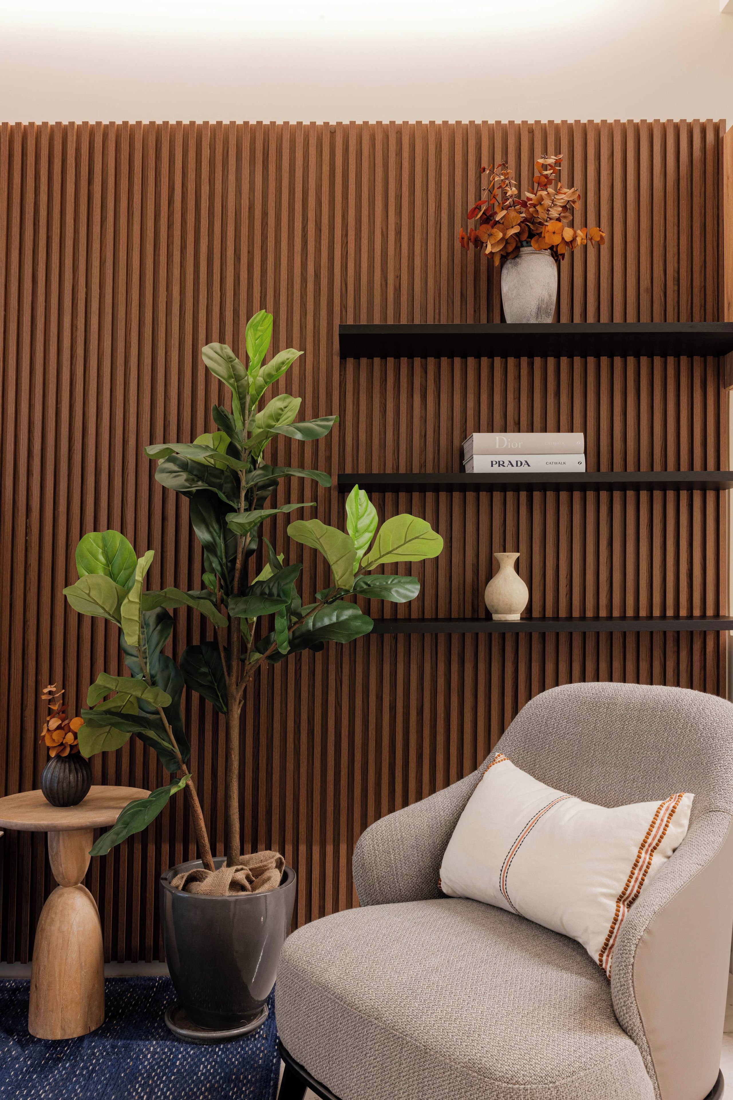 Une pièce avec un mur recouvert de panneaux de bois verticaux, une plante en pot, une chaise avec un coussin et une étagère contenant des livres et un vase.