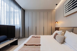 Moderni hotellihuone, jossa on suuri sänky, televisio ja näkymä kaupunkiin ohuiden verhojen läpi.