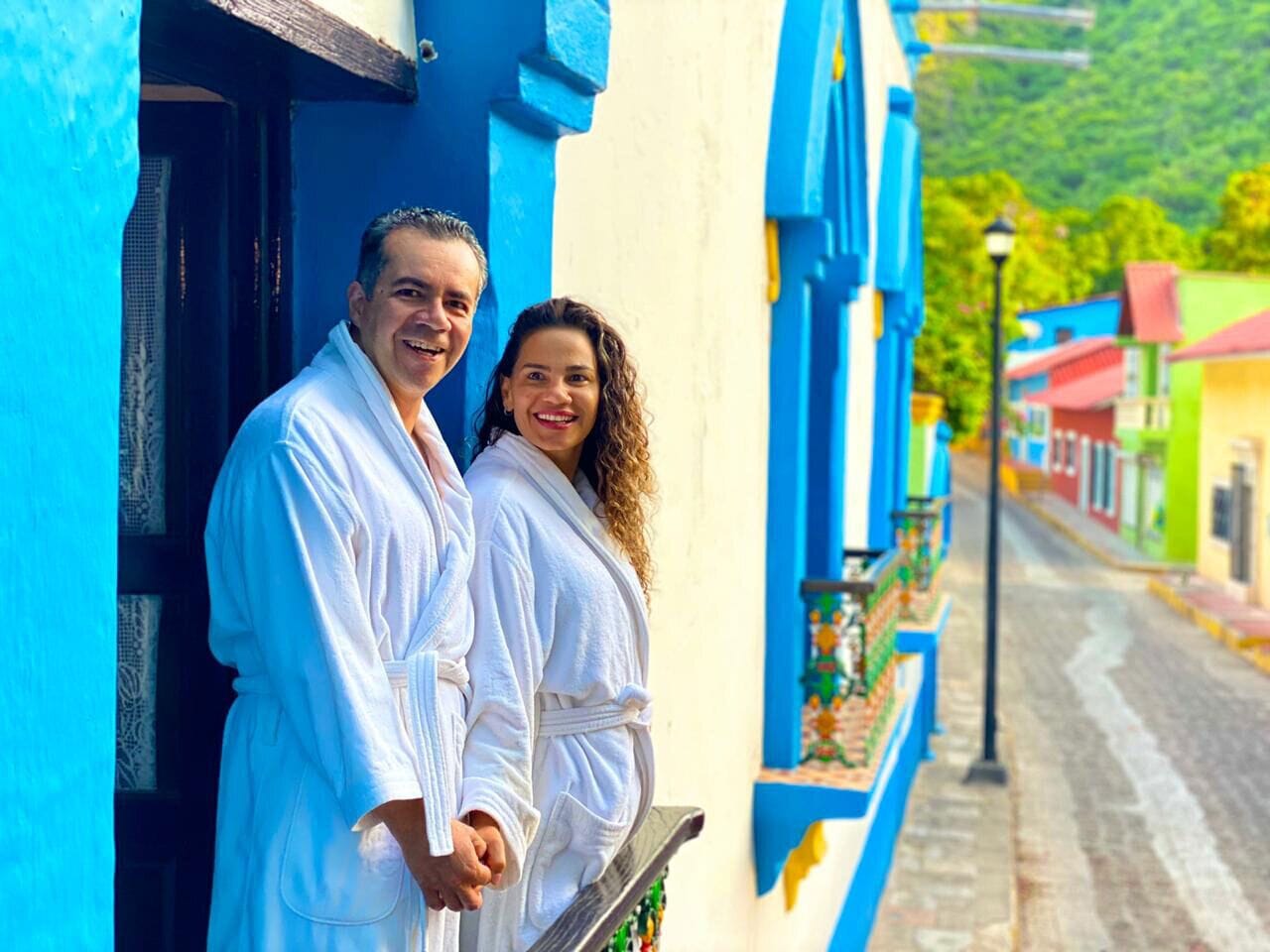 Una pareja parada en un balcón con vista a la calle, edificios pintados de azul y amarillo y una montaña al fondo.