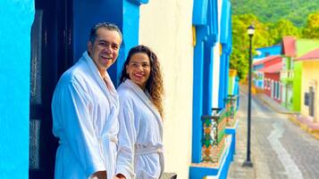 Una pareja parada en un balcón con vista a la calle, edificios pintados de azul y amarillo y una montaña al fondo.