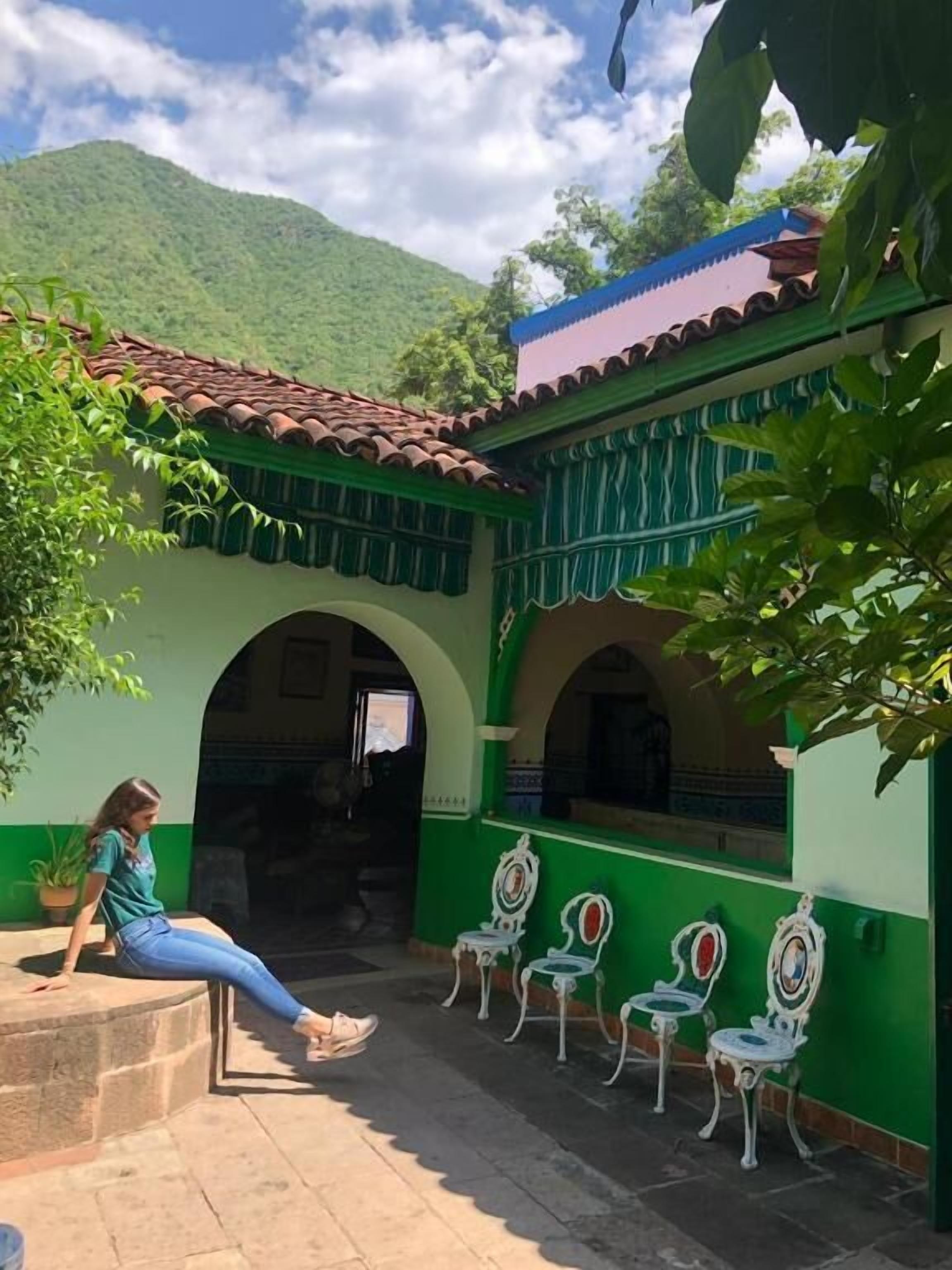 A courtyard with a green building, a woman sitting on a bench, and white chairs.