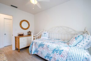 A bedroom with a white bed, a wooden dresser, a round mirror, and a ceiling fan. - Coastal Cocoanut Cottage ~ Whitaker Bayou~Bay (Sarasota)