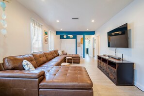 A spacious living area with a large brown leather sectional sofa, a flat-screen TV mounted on the wall, and a wooden entertainment unit. - Coastal Cocoanut Cottage ~ Whitaker Bayou~Bay (Sarasota)