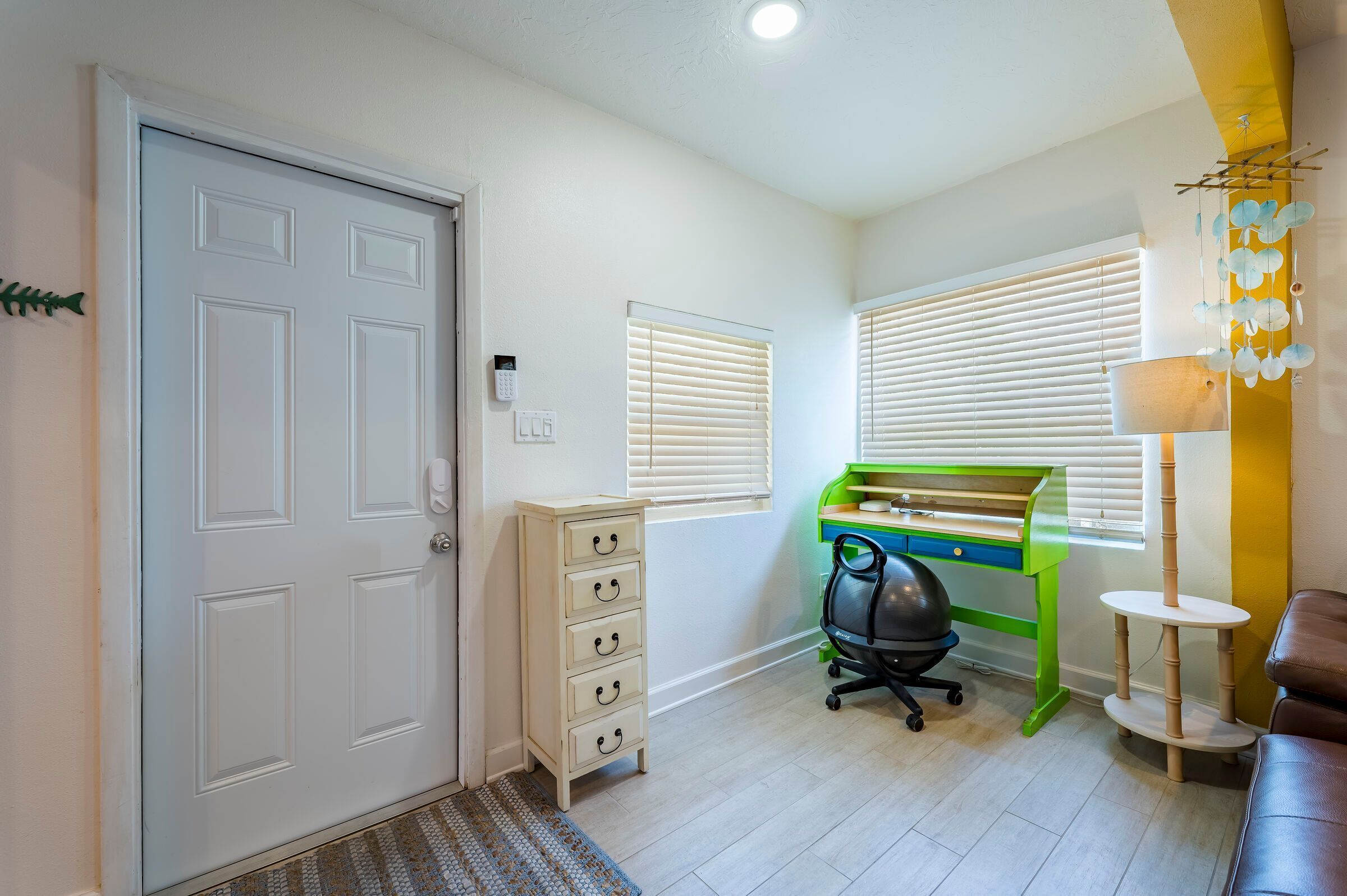 A room with a door, a desk with drawers, a chair, a lamp, and a window with blinds.