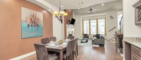 A modern dining area with a large table set for four, a chandelier, and a painting on the wall.