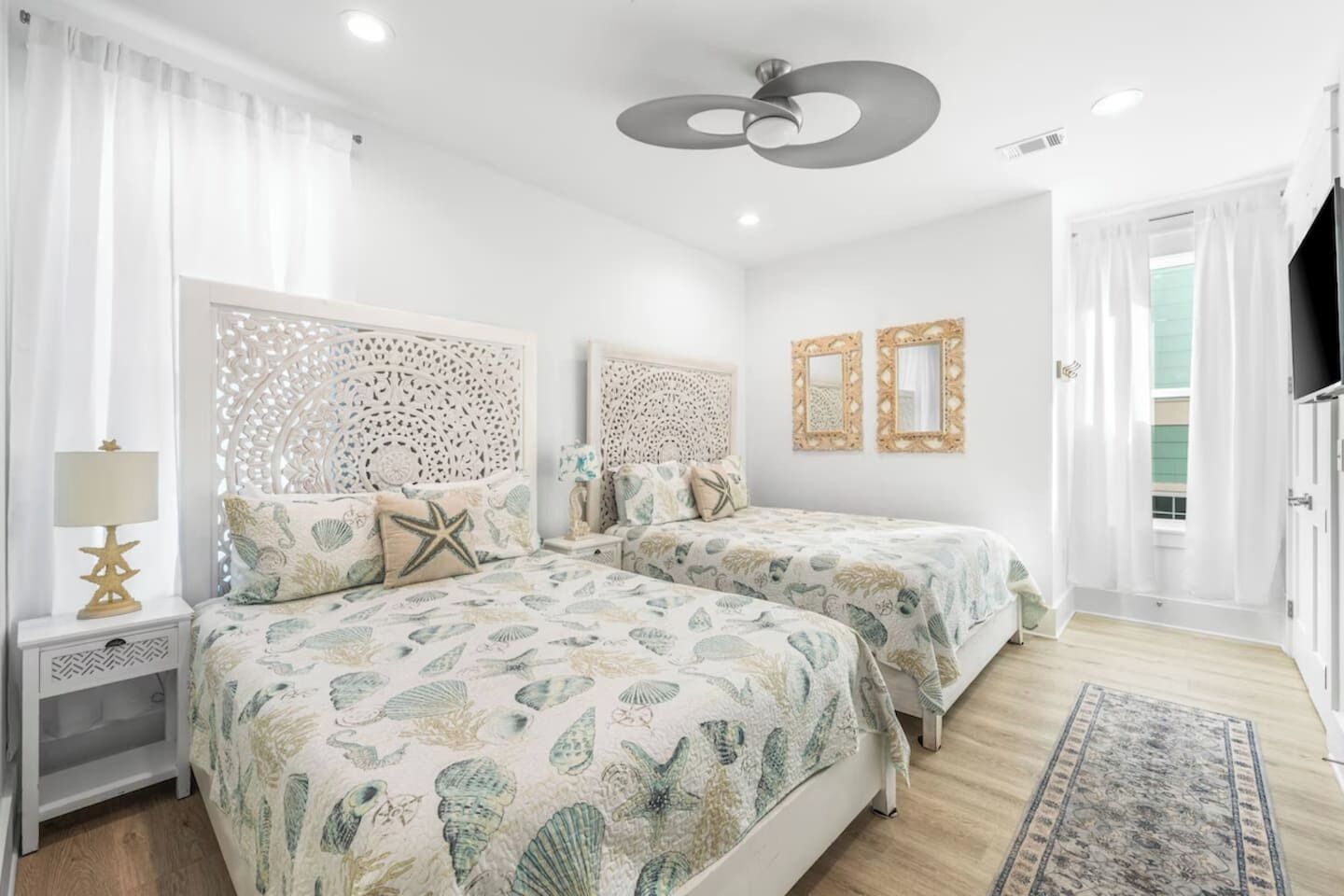 A bedroom with two beds, a nightstand with a lamp, a ceiling fan, and a view of the outside.