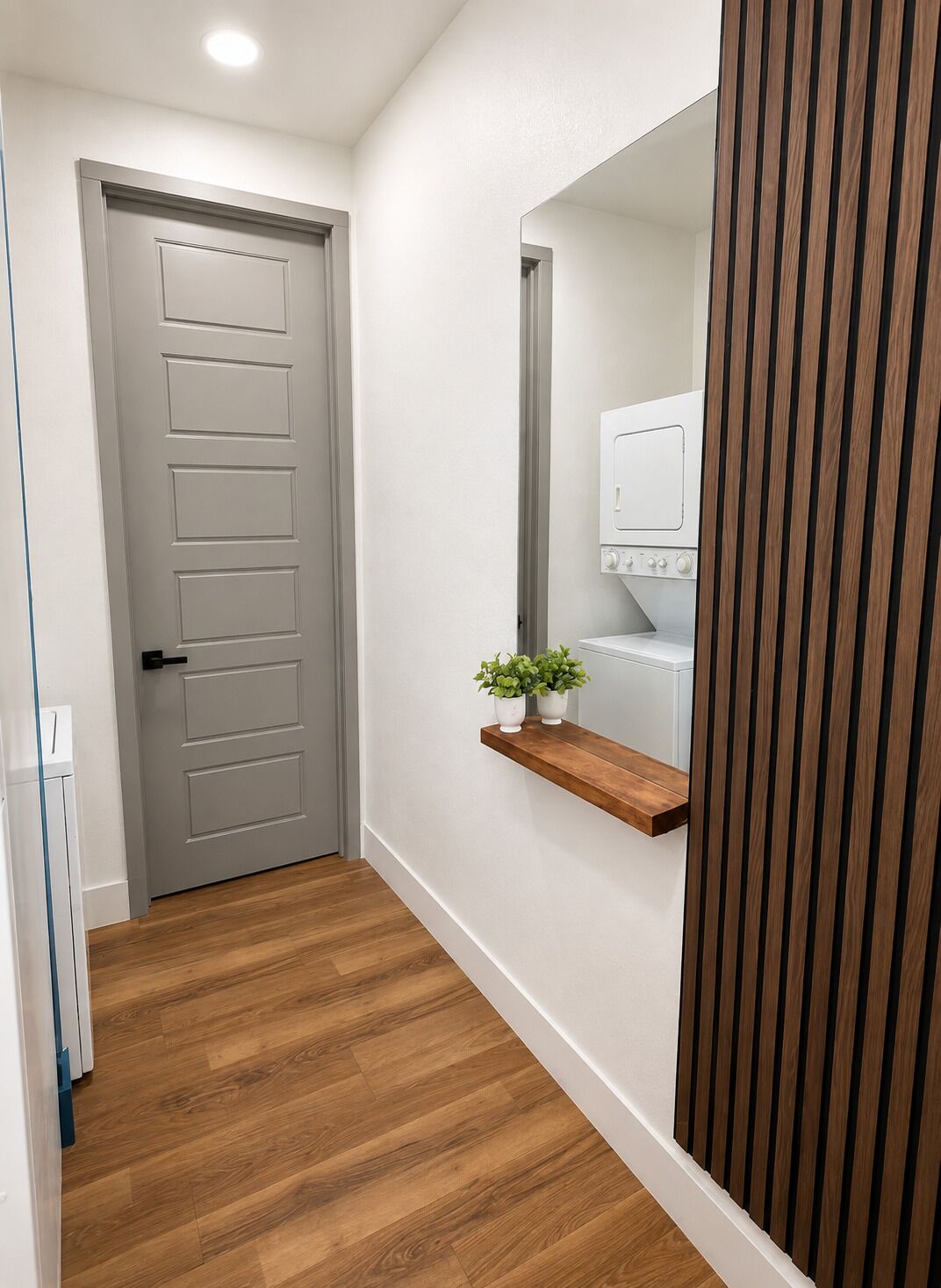 A hallway with a wooden floor, a grey door, and a white wall with a mirror and a shelf holding two potted plants.