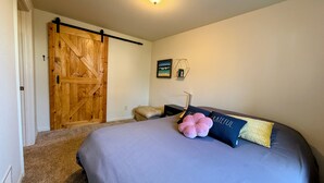 A bedroom with a large bed, a wooden door, a small stool, and a wall-mounted light fixture. - Gather in the Saddle - Spacious 3BR (Cheyenne)