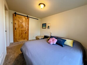 A bedroom with a large bed, a wooden door, a small stool, and a wall-mounted light fixture. - Gather in the Saddle - Spacious 3BR (Cheyenne)