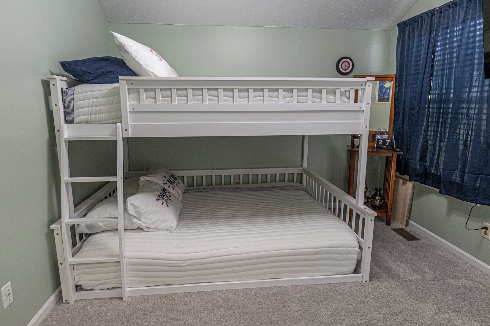 A bunk bed with a lower bunk, a bedside table with a clock, and a window with curtains.