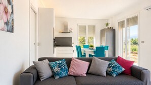 A modern living room with a grey sofa, teal and pink cushions, a white dining area with blue chairs, and a kitchen with a grey range hood. - Un Nido sul Garda by Case al Lago (Lonato)