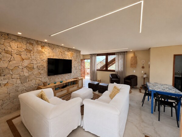 A modern living room with a stone wall, a flat-screen TV, white sofas, a wooden console table, and a dining area with a blue table and black chairs.