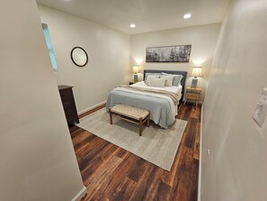 Un dormitorio con cama, lámparas de noche, un banco, cómoda y un cuadro en la pared.