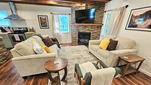 A living room with two sofas, a coffee table, a fireplace, a TV, and a kitchen in the background. - Charming, streamside, 3-bedroom country house in Confluence! (Confluence)