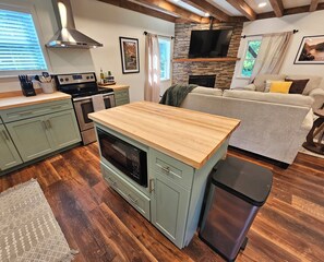 Cocina compacta con una isla central, horno integrado y estufa. Hay una chimenea de piedra, un televisor montado encima y un sofá en la sala.
