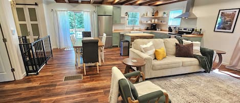 A spacious living area with a wooden floor, a sofa, a dining table, and a kitchen in the background.