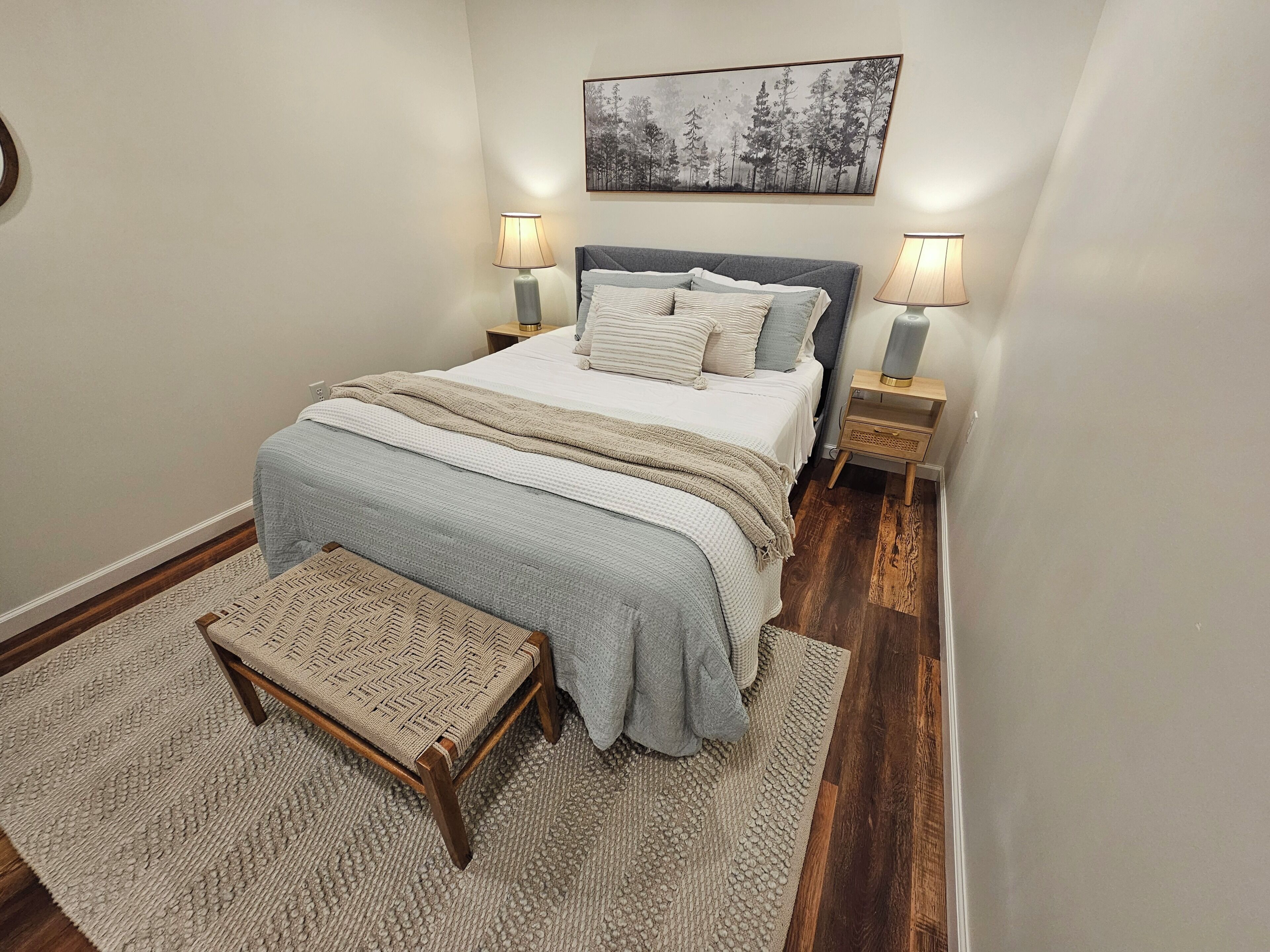 A bedroom with a bed, bedside lamps, a rug, a bench, and a wall-mounted picture.