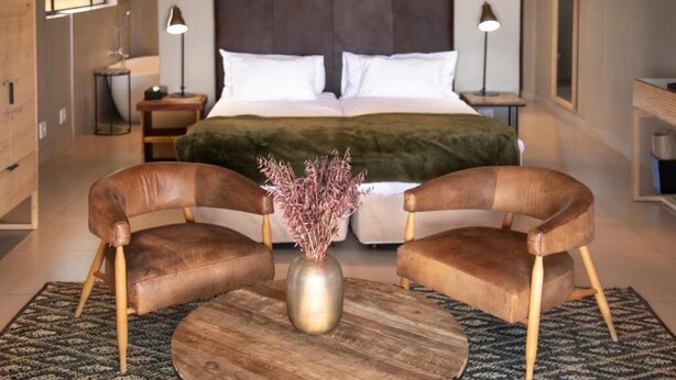 Un dormitorio con una cama grande, dos sillas, una mesa redonda y un jarrón con flores secas.