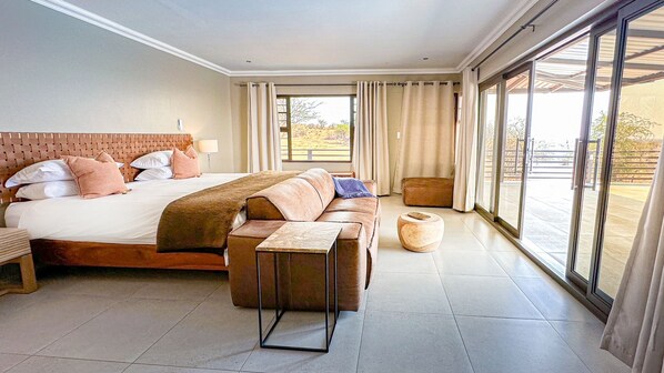 A hotel room with a large bed, a sofa, a small table, and a view through large windows.