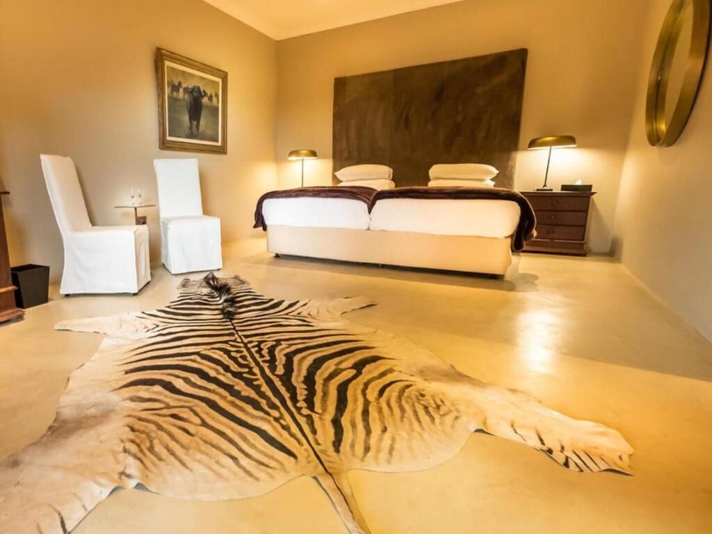 A bedroom with a large bed, two bedside tables, a chair, and a zebra hide on the floor.