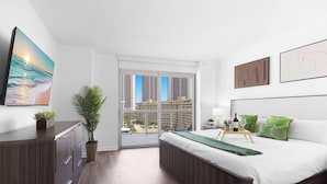 Quarto moderno com cama, mesa de cabeceira, luminária, planta em vaso e vista da cidade pela janela.