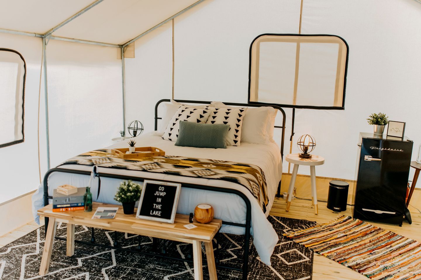 Una habitación con carpa, una cama, una mesita, un refrigerador negro y una alfombra estampada.