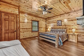 Ein Schlafzimmer in einer Blockhütte mit Bett, Nachttisch, Deckenventilator und einem Fenster mit Jalousien.