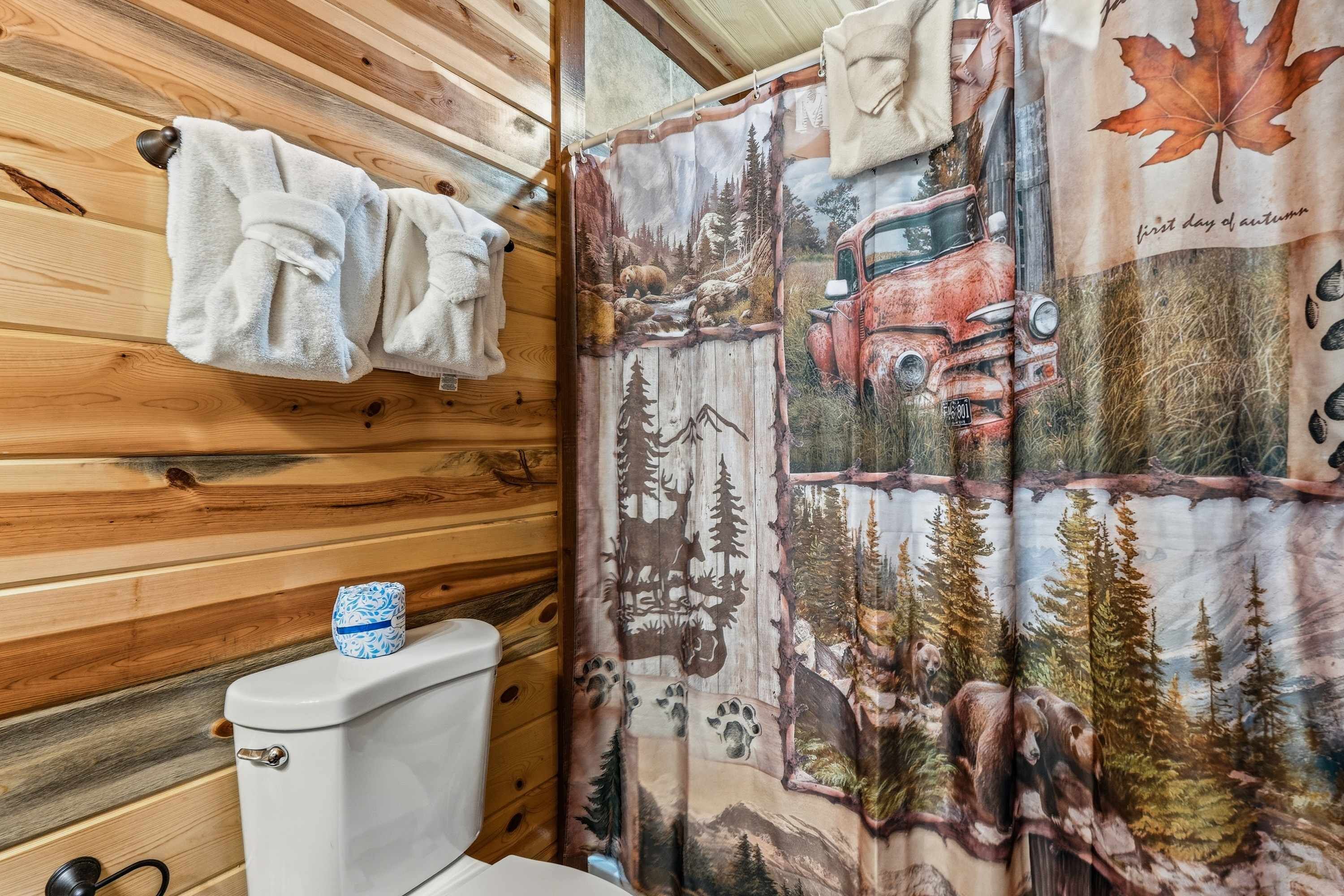 A bathroom with wooden walls, a white toilet, and a shower curtain with a forest and vehicle design.