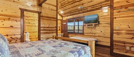 Interior de una cabaña de madera con una cama, una ventana con vistas y un televisor de pared.