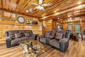 Ein Interieur einer Blockhütte mit zwei Sofas, einem Deckenventilator, einem Holz-Couchtisch und einem Küchenbereich im Hintergrund.