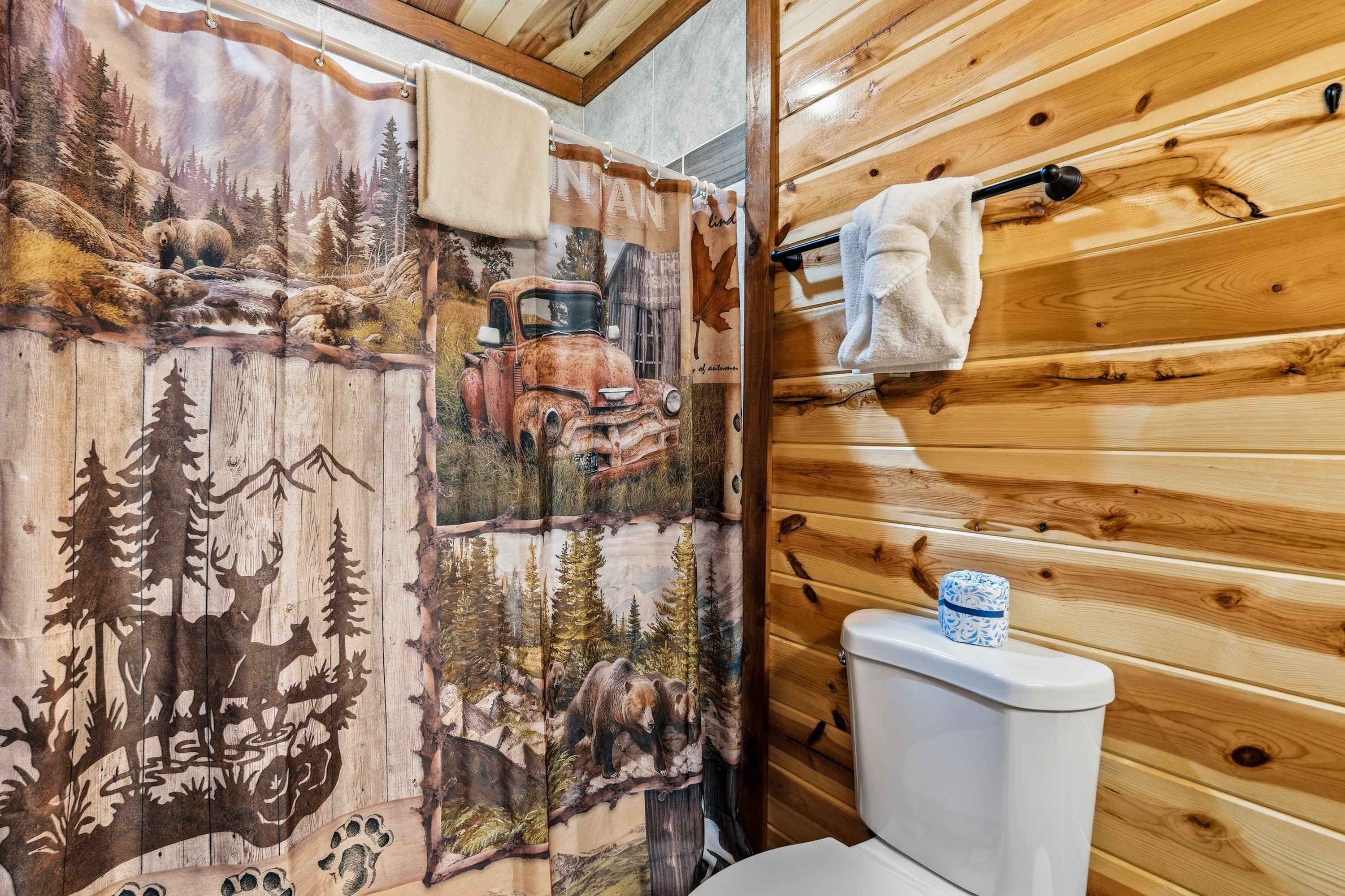 Un baño con detalles de madera, una cortina de ducha con diseño de bosque y vehículo, y un inodoro con un pequeño adorno encima.