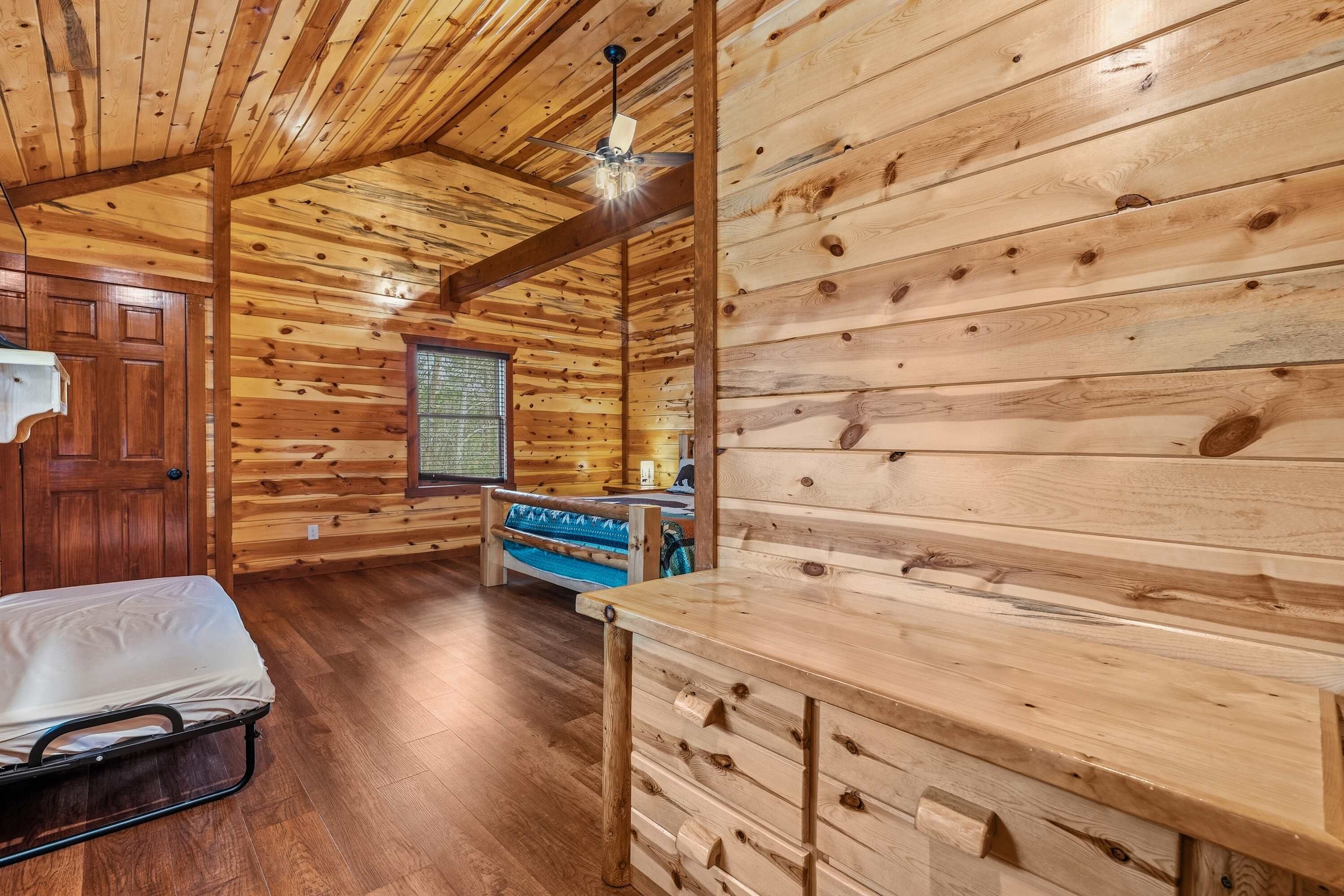 A wooden cabin interior with a bed, a bench, a window with a view, and a ceiling fan.