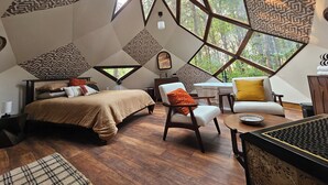 Quarto com design geométrico no teto, uma cama, uma cadeira, uma mesa e vista para árvores através de grandes janelas.